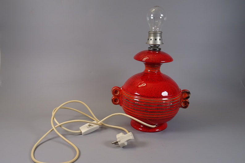 May include: A red ceramic table lamp with a bulb. The lamp has a round base and body with horizontal lines and two small handles. The lamp is connected to a white cord with a switch and plug.