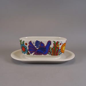 Könnte beinhalten: Ein weißes Keramik-Serviergeschirr mit passender Untertasse. Das rechteckige Geschirr zeigt ein farbenfrohes Blumen- und Vogelmuster in Blau, Orange, Rot und Grün. Das Set ist zum Servieren von Speisen gedacht.