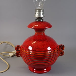 May include: A bright red ceramic table lamp with a bulb. The lamp has a round base and body, with horizontal stripes and two sets of decorative handles. The lamp is connected to a cream-coloured electrical cord.