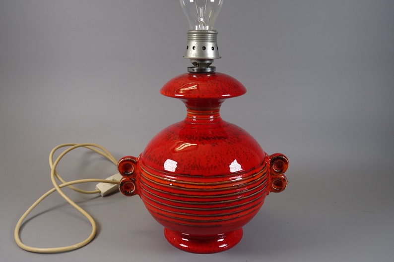 May include: A bright red ceramic table lamp with a bulb. The lamp has a round base and body, with horizontal stripes and two sets of decorative handles. The lamp is connected to a cream-coloured electrical cord.