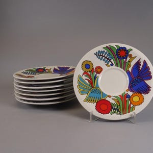 Könnte beinhalten: Ein Satz weißer Keramikuntertassen mit einem farbenfrohen Vogel- und Blumendesign. Die Untertassen haben eine zentrale Vertiefung für eine Tasse und sind mit blauen, gelben, roten und grünen Elementen verziert. Ein Stapel Untertassen ist sichtbar.