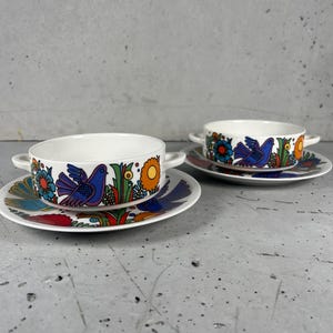 Könnte beinhalten: Zwei weiße Keramik-Suppenschalen mit Henkeln, jeweils auf einer passenden Untertasse. Die Schalen und Untertassen weisen ein farbenfrohes Design aus blauen Vögeln, Blumen und Blättern auf. Das Design umfasst Orange-, Blau- und Gelbtöne.