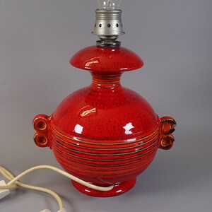 May include: A red ceramic table lamp with a textured finish. The lamp features a bulb holder at the top and a rounded base. The lamp has a distinctive design with horizontal stripes and decorative handles. A white electrical cord with a switch is attached.