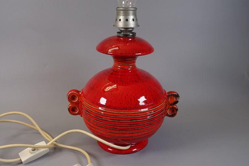 May include: A red ceramic table lamp with a textured finish. The lamp features a bulb holder at the top and a rounded base. The lamp has a distinctive design with horizontal stripes and decorative handles. A white electrical cord with a switch is attached.