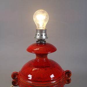 May include: A red ceramic table lamp with a lit bulb. The lamp features a bulb socket, a mushroom-shaped top, and a rounded base with decorative handles. The lamp's surface has a speckled texture and horizontal lines.