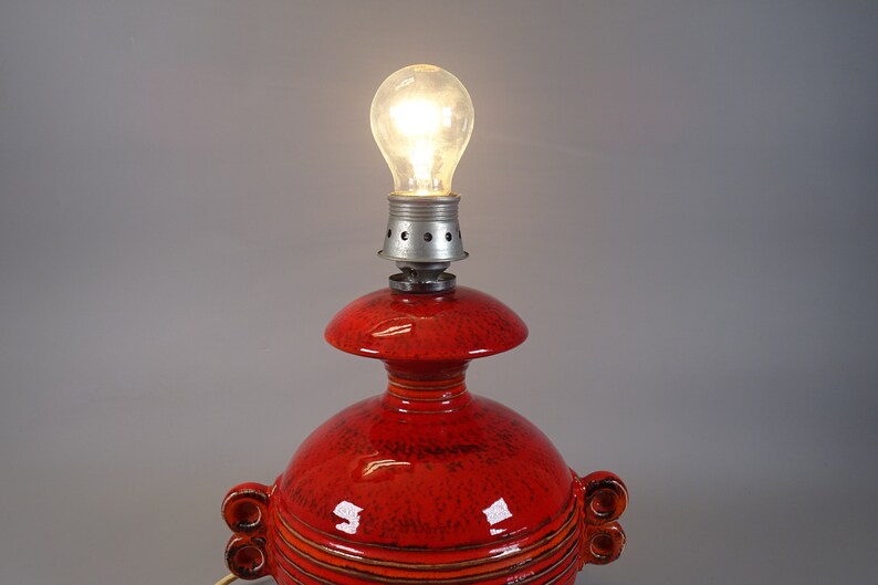 May include: A red ceramic table lamp with a lit bulb. The lamp features a bulb socket, a mushroom-shaped top, and a rounded base with decorative handles. The lamp's surface has a speckled texture and horizontal lines.