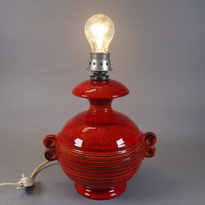 May include: A red ceramic table lamp with a bulb. The lamp has a round base and body with horizontal stripes and two sets of decorative handles. The lamp's top has a smaller, flared shape. The bulb is lit.