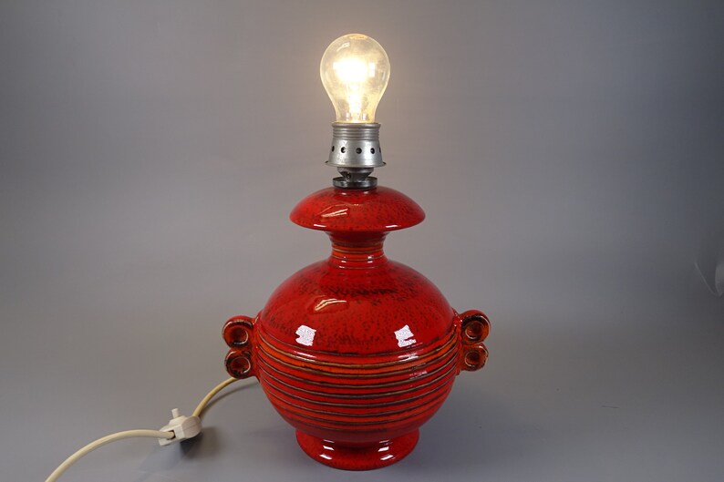 May include: A red ceramic table lamp with a bulb. The lamp has a round base and body with horizontal stripes and two sets of decorative handles. The lamp's top has a smaller, flared shape. The bulb is lit.