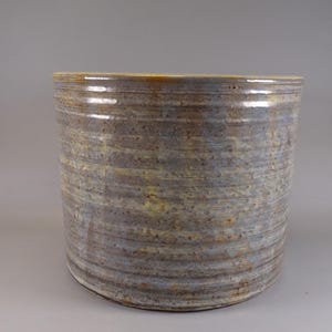 Può includere: Un vaso cilindrico in ceramica con superficie strutturata. Il vaso ha una palette di colori neutri con sfumature di grigio, marrone e giallo. Il design presenta linee orizzontali, creando un'estetica rustica.