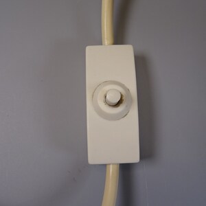 May include: Close-up of a white light switch with a rounded button, attached to a cream-coloured cord. The switch is rectangular and mounted on a grey wall. The button is slightly discoloured.