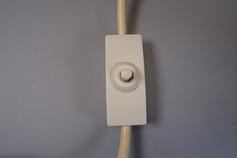 May include: Close-up of a white light switch with a rounded button, attached to a cream-coloured cord. The switch is rectangular and mounted on a grey wall. The button is slightly discoloured.