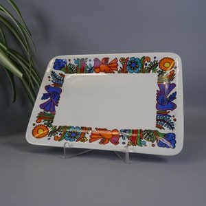 Könnte beinhalten: Rechteckige weiße Keramik-Servierplatte mit einem farbenfrohen Rand mit Vögeln, Blumen und Blättern in Orange, Blau und Grün. Die Platte wird auf einem transparenten Ständer präsentiert.