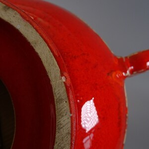 May include: Close-up of a bright red ceramic vase with a glossy finish. The vase has a rounded body and a small spout. The base of the vase is a lighter, unglazed colour. The overall design is simple and modern.