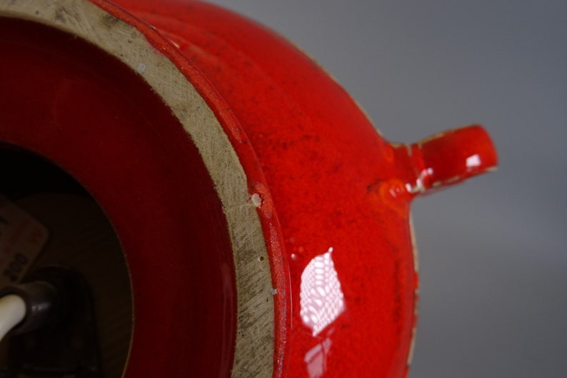 May include: Close-up of a bright red ceramic vase with a glossy finish. The vase has a rounded body and a small spout. The base of the vase is a lighter, unglazed colour. The overall design is simple and modern.
