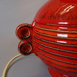 May include: A bright red ceramic lamp with horizontal brown and orange stripes. The lamp has two decorative, circular handles with a white and black trim. A cream-coloured cord extends from the base.