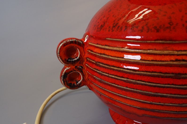 May include: A bright red ceramic lamp with horizontal brown and orange stripes. The lamp has two decorative, circular handles with a white and black trim. A cream-coloured cord extends from the base.