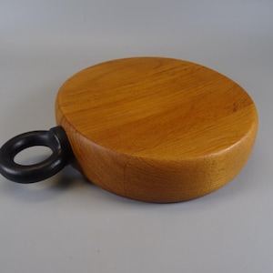 Puede incluir: Una tabla de cortar redonda de madera con un asa gris oscuro. La tabla es de madera marrón claro y tiene una superficie lisa. El asa es un anillo circular unido al lateral de la tabla.