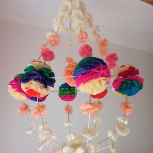 Puede incluir: Una lámpara de araña de flores de papel de colores con flores de papel blancas, naranjas, rosas, verdes, azules y rojas. La lámpara de araña está hecha de flores de papel enhebradas en delgadas varillas de madera.