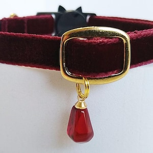 Pode incluir: Uma coleira de gato de veludo bordô com uma fivela dourada e um pequeno pingente vermelho em forma de gota. A coleira tem um fecho preto em forma de gato. A fivela e o pingente dourados adicionam um toque de elegância a este acessório para animais de estimação.