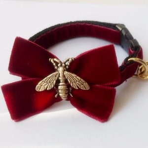 May include: A burgundy velvet bow tie with a gold bee charm. The bow tie is attached to a black adjustable collar.