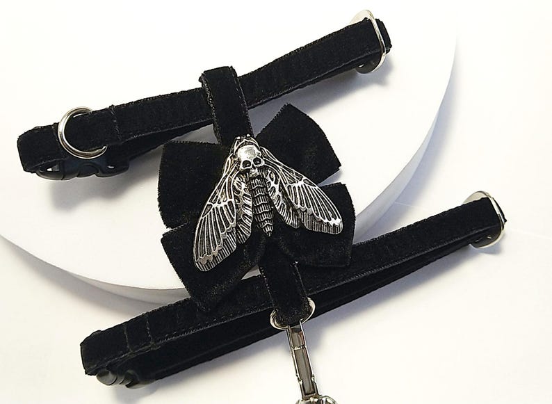 May include: Black velvet choker with a silver-toned moth pendant featuring a skull design. The choker has a bow detail and adjustable straps with metal hardware. A gothic-inspired accessory.