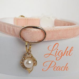 May include: A light peach velvet cat collar with a gold buckle and a gold charm with a pearl and rhinestones. The charm is shaped like a heart. The text "Light Peach" is written below the collar.