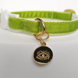 May include: A lime green velvet cat collar with a white buckle. The collar features a gold-colored charm with a black background and an eye design with a small crystal in the center. The collar is designed for pets.