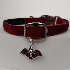 Puede incluir: Un collar de terciopelo burdeos para mascotas con una hebilla plateada y un dije de murci&eacute;lago. El dije de murci&eacute;lago tiene detalles de esmalte rojo y negro. El collar es de color rojo oscuro.