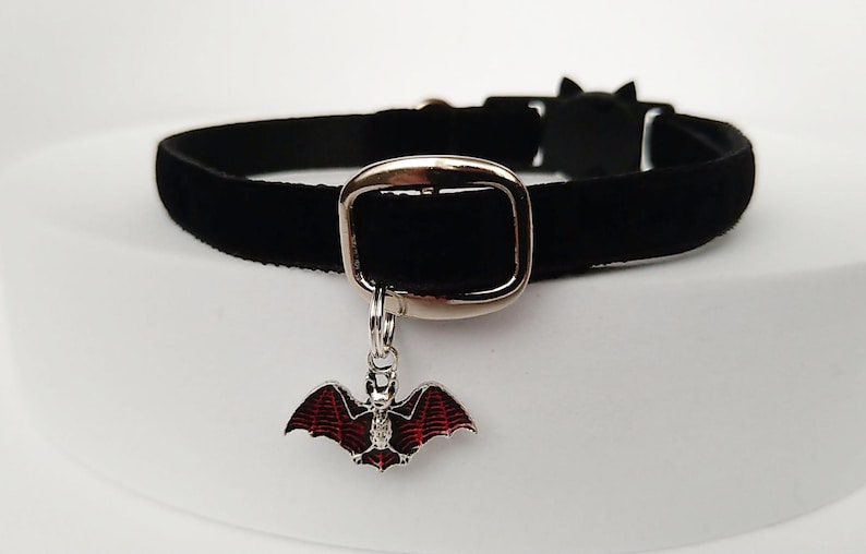 Puede incluir: Un collar gargantilla de terciopelo negro con una hebilla plateada y un dije de murci&eacute;lago. El dije de murci&eacute;lago tiene detalles de esmalte rojo y negro. El collar se muestra sobre una superficie blanca.