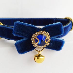 Peut inclure: Un collier pour chat en velours bleu marine avec un nœud, une clochette dorée et un ornement central incrusté de bijoux. Le collier a une boucle noire et des éléments dorés.