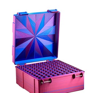 May include: Open, pink and blue storage case with a geometric starburst design on the lid. The interior is filled with small, cylindrical compartments. The case has a hinged lid and metal latches.