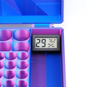 May include: A blue and purple 3D-printed storage box with a digital thermometer displaying 29% humidity and 76.2°F. The box has multiple compartments and a hinged lid. The interior is divided into sections.