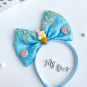 May include: A light blue headband with a large bow featuring an ice cream cone and lollipop accents. The bow is decorated with colorful sprinkles and the words "My Bows".