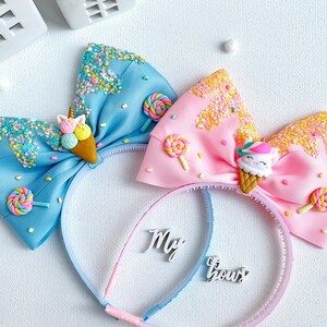 May include: Two headbands with large bows. One bow is light blue, the other is pink. Each bow is decorated with an ice cream cone, sprinkles, and a lollipop. The text "My Bow" is on the headband.