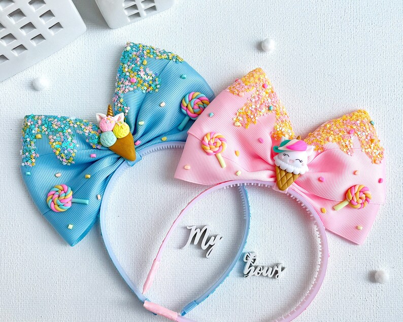 May include: Two headbands with large bows. One bow is light blue, the other is pink. Each bow is decorated with an ice cream cone, sprinkles, and a lollipop. The text "My Bow" is on the headband.