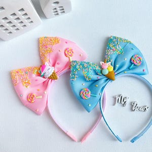 May include: Two colorful headbands with bow accents. One is pink, the other is blue. Each bow features a unicorn ice cream cone, sprinkles, and a lollipop. The headbands are decorated with glitter and are perfect for a child.
