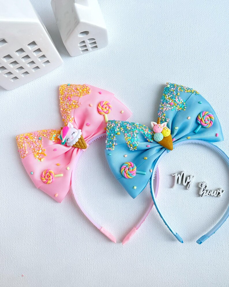 May include: Two colorful headbands with bow accents. One is pink, the other is blue. Each bow features a unicorn ice cream cone, sprinkles, and a lollipop. The headbands are decorated with glitter and are perfect for a child.