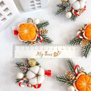 May include: A collection of festive hair clips featuring felted orange slices, cotton, pinecones, and candy canes. Each clip is accented with faux greenery, gold glitter, and pearls. The clips are attached to red ribbon bases. A ruler shows the clips are approximately 5 inches in length.