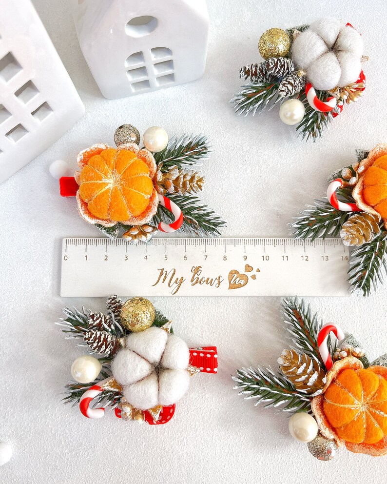 May include: A collection of festive hair clips featuring felted orange slices, cotton, pinecones, and candy canes. Each clip is accented with faux greenery, gold glitter, and pearls. The clips are attached to red ribbon bases. A ruler shows the clips are approximately 5 inches in length.