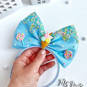 May include: A light blue headband with a large bow featuring a whimsical ice cream cone and lollipop design. The bow is decorated with colorful sprinkles and glitter, and the headband is held by a hand. The text "My Bows" is visible.