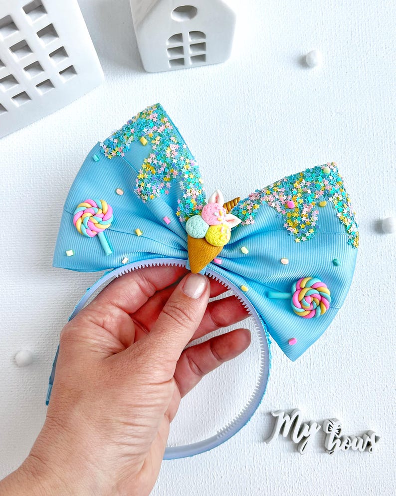 May include: A light blue headband with a large bow featuring a whimsical ice cream cone and lollipop design. The bow is decorated with colorful sprinkles and glitter, and the headband is held by a hand. The text "My Bows" is visible.