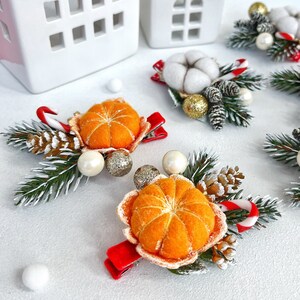 May include: Christmas-themed hair clips featuring felt mandarin oranges, pinecones, and candy canes. The clips have red velvet bows and are accented with faux pearls and glittery gold ornaments. White ceramic houses are in the background.