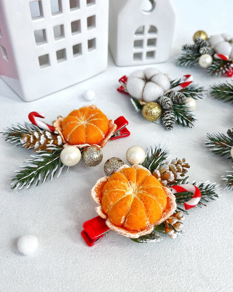 May include: Christmas-themed hair clips featuring felt mandarin oranges, pinecones, and candy canes. The clips have red velvet bows and are accented with faux pearls and glittery gold ornaments. White ceramic houses are in the background.