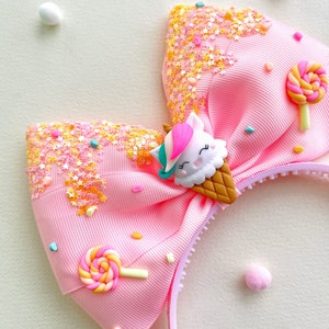 May include: Pink headband with a large bow decorated with a unicorn ice cream cone, lollipop, and star-shaped sprinkles. The bow is made of grosgrain ribbon. The headband is a fun accessory for children.