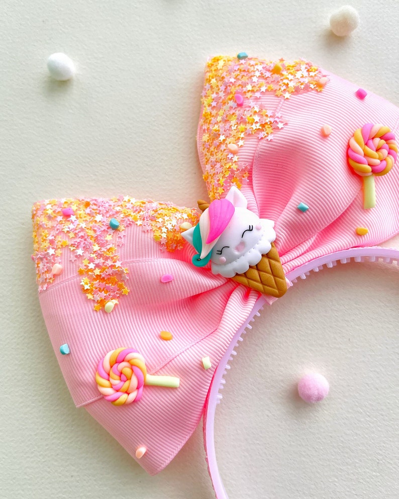 May include: Pink headband with a large bow decorated with a unicorn ice cream cone, lollipop, and star-shaped sprinkles. The bow is made of grosgrain ribbon. The headband is a fun accessory for children.