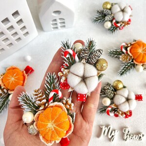 May include: A collection of Christmas-themed hair clips. Each clip features a felt cotton flower, miniature pine cones, candy canes, and orange slices. The clips are adorned with faux pearls and glittery accents, set against a backdrop of white and green.