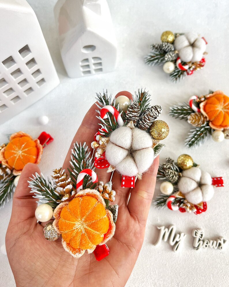 May include: A collection of Christmas-themed hair clips. Each clip features a felt cotton flower, miniature pine cones, candy canes, and orange slices. The clips are adorned with faux pearls and glittery accents, set against a backdrop of white and green.