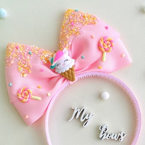 May include: A pink headband with a large bow featuring a unicorn ice cream cone and lollipop accents. The bow has glitter and star details. The words "My bows" are on the headband.