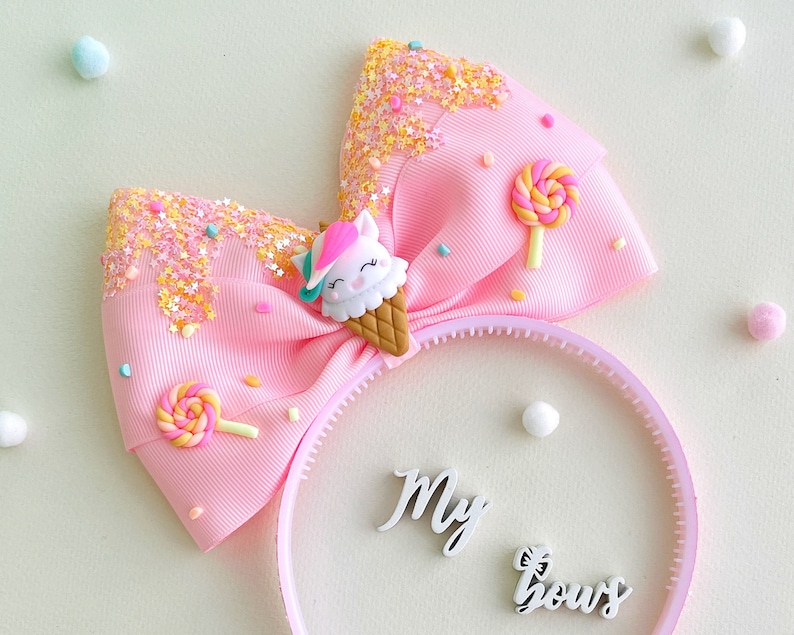 May include: A pink headband with a large bow featuring a unicorn ice cream cone and lollipop accents. The bow has glitter and star details. The words "My bows" are on the headband.