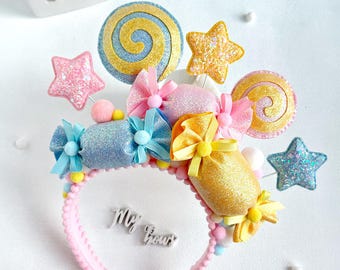Könnte beinhalten: Ein rosafarbenes Stirnband, verziert mit glitzernden bonbonförmigen Ornamenten, spiralförmigen Lollis und Sternen in Blau-, Gelb- und Rosatönen. Das Stirnband ist mit kleinen Pompons und Schleifen versehen, der Text My Bows ist mittig sichtbar.